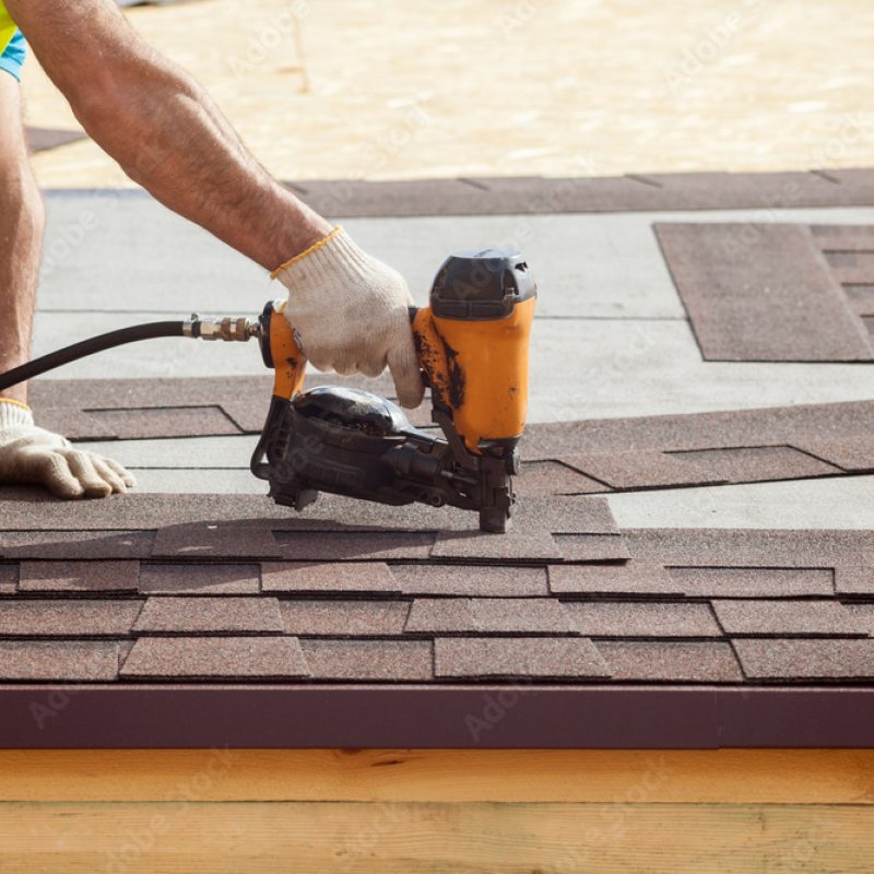 Construction worker putting the asphalt roofing (shingles) with nail gun on a new frame house